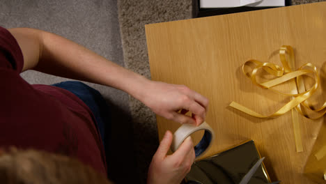 Overhead-Shot-Of-Man-Using-Sticky-Tape-And-Ribbon-To-Gift-Wrapped-Present-On-Table-At-Home